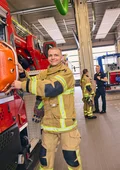 Feuerwehrmann in Schutzkleidung an einem roten Feuerwehrfahrzeug in einer Fahrzeughalle, im Hintergrund sind zwei weitere Personen in Feuerwehr- und Einsatzkleidung im Gespräch.
