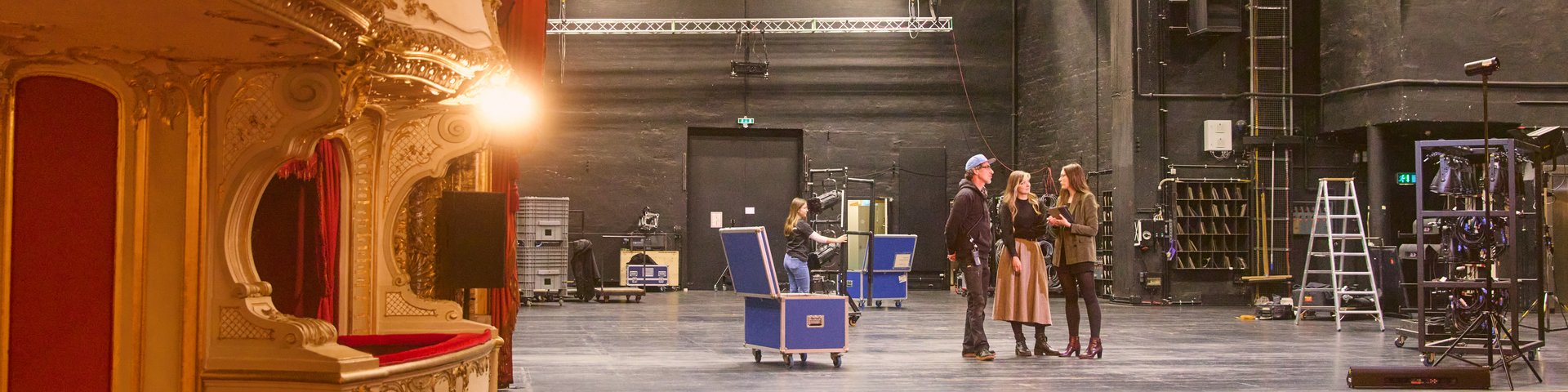 Bühnenraum eines Theaters mit rotem Vorhang, goldverzierten Logen, roten Sitzreihen und drei Personen auf der Bühne mit technischer Ausstattung.