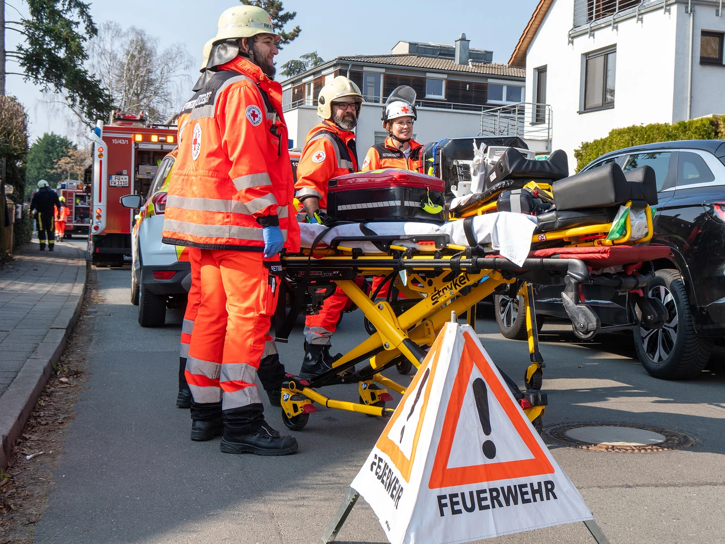 Rettungskräfte in orangefarbener Schutzkleidung mit Helmen neben einer gelben Trage und einem Warnschild mit der Aufschrift "FEUERWEHR".