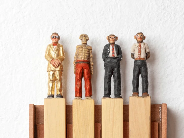 Vier kleine Holzfiguren mit Affengesichtern in unterschiedlichen Anzügen stehen auf vier Holzklötzen in einem Holzrahmen an einer weißen Wand. Klick öffnet Veranstaltungstipp.