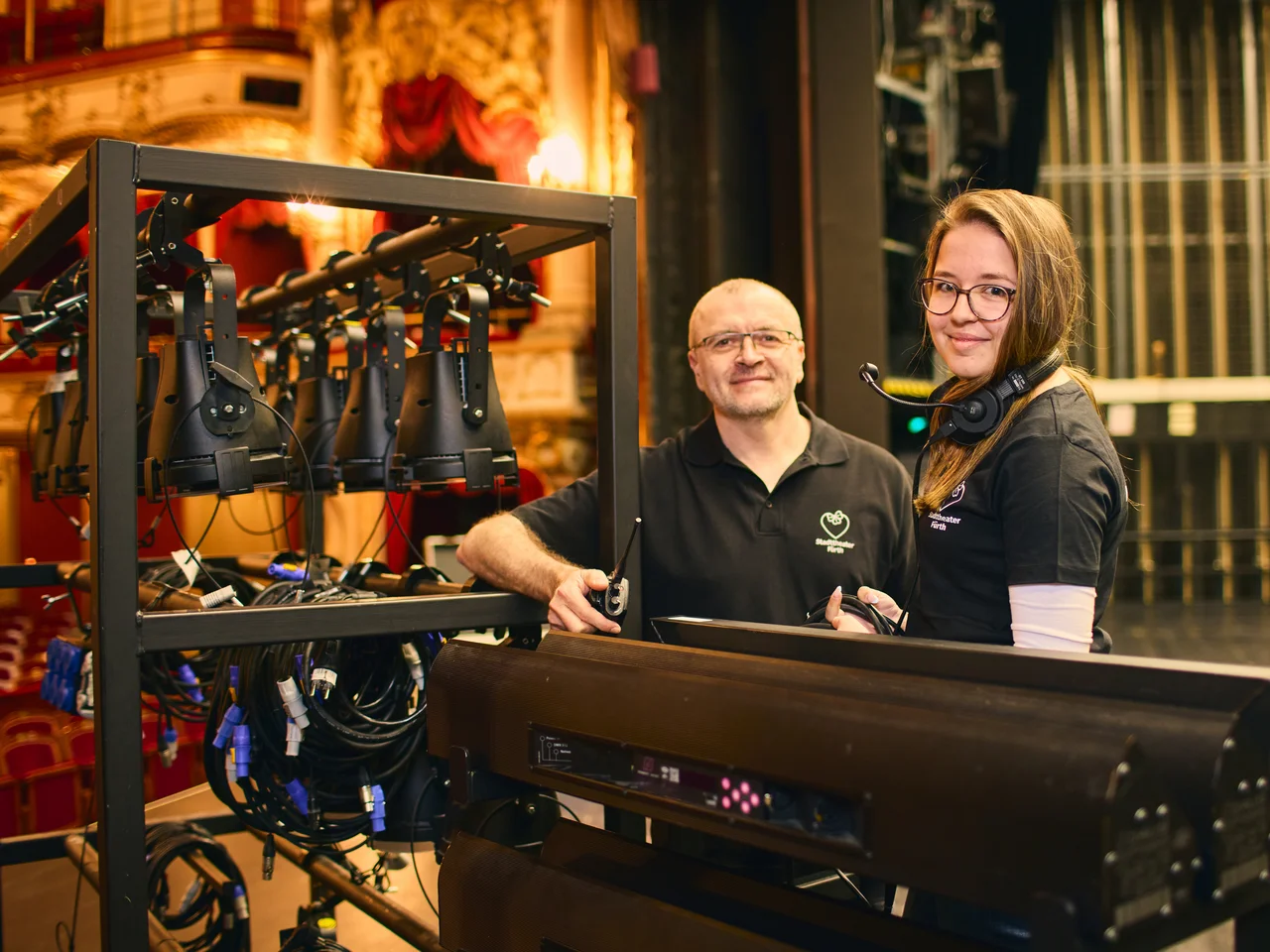 Zwei Personen stehen an einem großen technischen Gerät mit mehreren Kabeln und Halterungen in einem Theater.