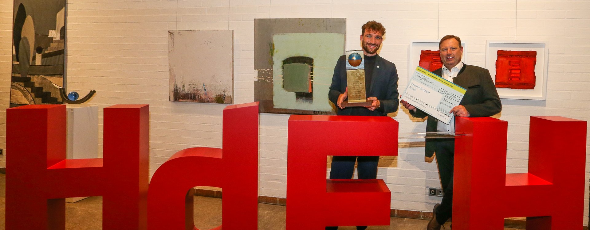 Zwei Männer mit Preis und Scheck in der Hand stehen hinter den roten Buchstaben HdFH, kurz für Hauptstadt des Fairen Handels.