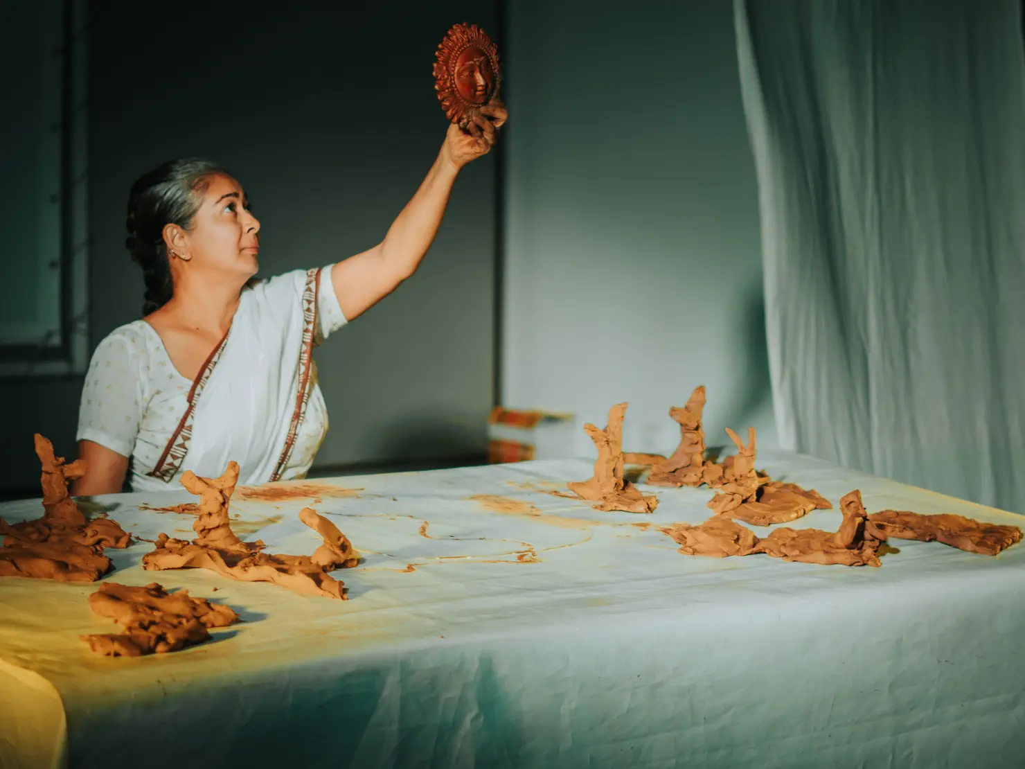 Eine Frau in traditioneller Kleidung hält ein geformtes Tonobjekt hoch, während sie am Tisch mit weiteren Tonfiguren arbeitet. Die Szene hat eine sanfte Beleuchtung.