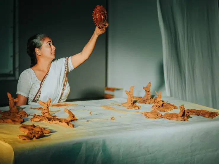 Eine Frau in traditioneller Kleidung hält ein geformtes Tonobjekt hoch, während sie am Tisch mit weiteren Tonfiguren arbeitet. Die Szene hat eine sanfte Beleuchtung.