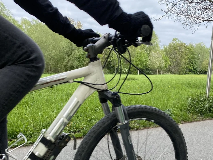 Nahaufnahme eines weißen Mountainbikes mit schwarzer Bereifung und einer Person in schwarzer Kleidung, die die Lenkergriffe hält, vor einer grünen Wiese und Bäumen