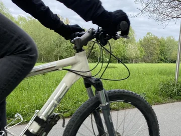 Nahaufnahme eines weißen Mountainbikes mit schwarzer Bereifung und einer Person in schwarzer Kleidung, die die Lenkergriffe hält, vor einer grünen Wiese und Bäumen