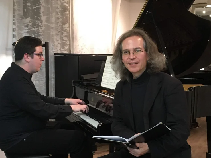 Zwei Männer in schwarzer Kleidung vor einem Flügel, einer spielt Klavier, der andere hält ein Notenheft und schaut in die Kamera