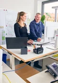 Zwei Personen stehen an einem Schreibtisch und blicken auf einen Computerbildschirm in einem hellen Büro.