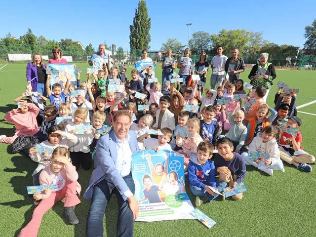 Jubelnde Kinder und gennante Personen sitzen und stehen im Kreis auf Sportplatz. Klick öffnet die Meldung.