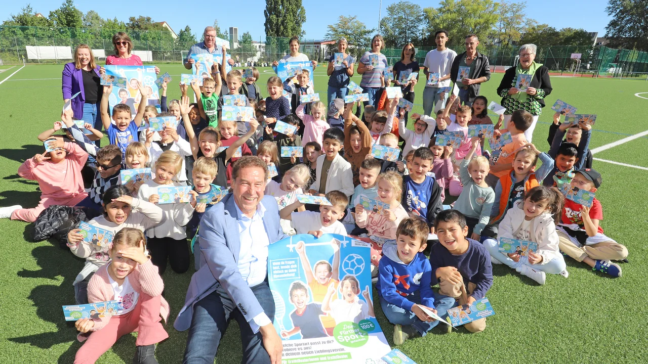 Jubelnde Kinder und gennante Personen sitzen und stehen im Kreis auf Sportplatz.
