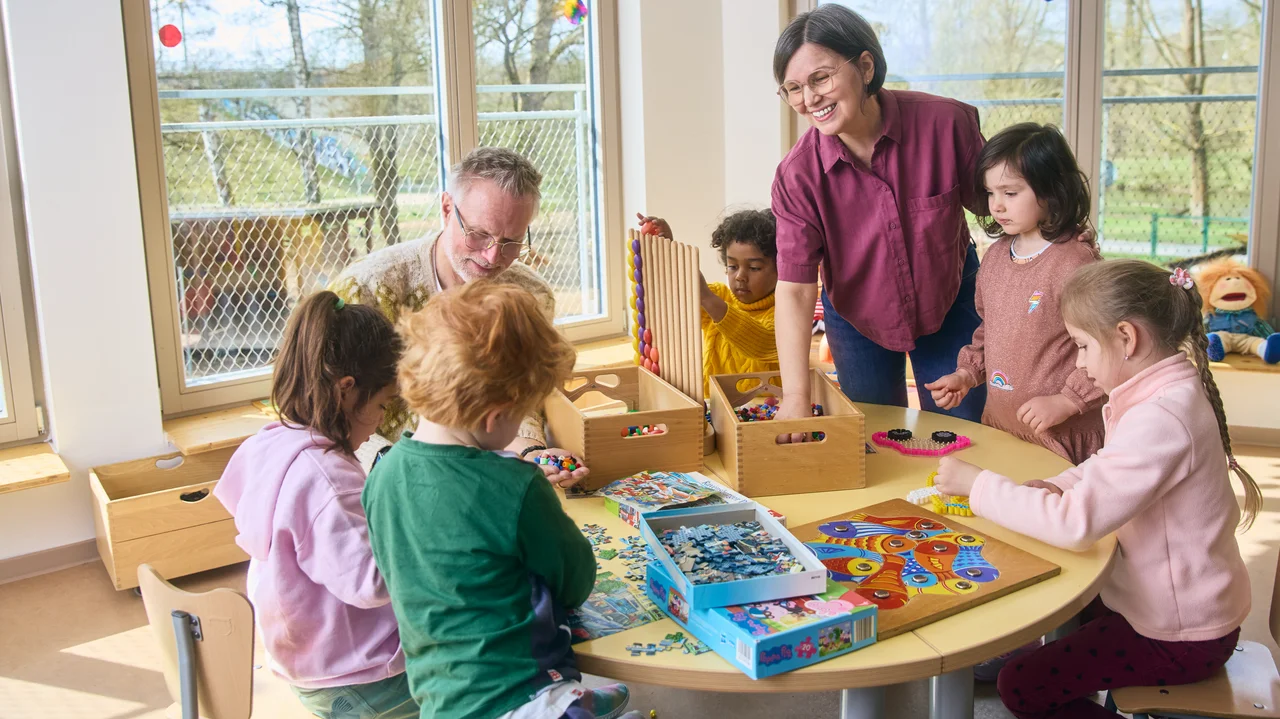 Kinder sitzen an einem runden Tisch und spielen mit verschiedenen Spielen und Puzzles, zwei erwachsene Personen stehen dabei.