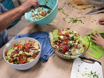 Zwei Schüsseln mit gemischtem Salat auf einem Holztisch, daneben Schneidebrett und Hände von zwei Personen.