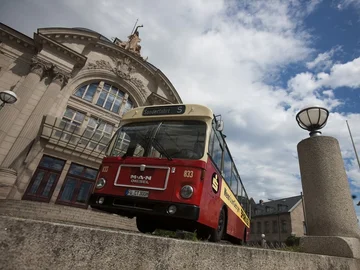 Alter roter Bus mit der Aufschrift 'Sonderfahrt' steht vor einem historischen Gebäude mit großen Fenstern und Säulen. Im Vordergrund sind zwei runde Straßenlaternen auf Betonpfeilern zu sehen.
