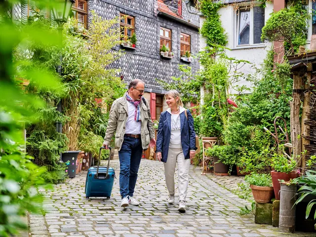 Ein Mann und eine Frau laufen eine Straße entlang und blicken sich gegenseitig an. Der Mann hat einen Koffer in der Hand. Klick öffnet die Meldung.