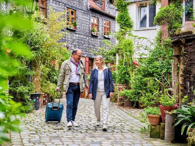 Ein Mann und eine Frau laufen eine Straße entlang und blicken sich gegenseitig an. Der Mann hat einen Koffer in der Hand. Klick öffnet Veranstaltungstipp.