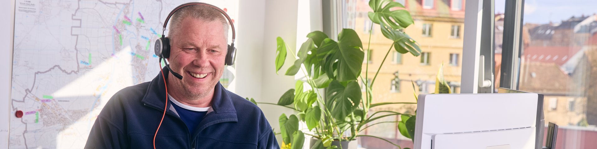 Mitarbeiter mit Headset sitzt am Schreibtisch vor einem Laptop und Monitor vor Fenster in hellem Büro.