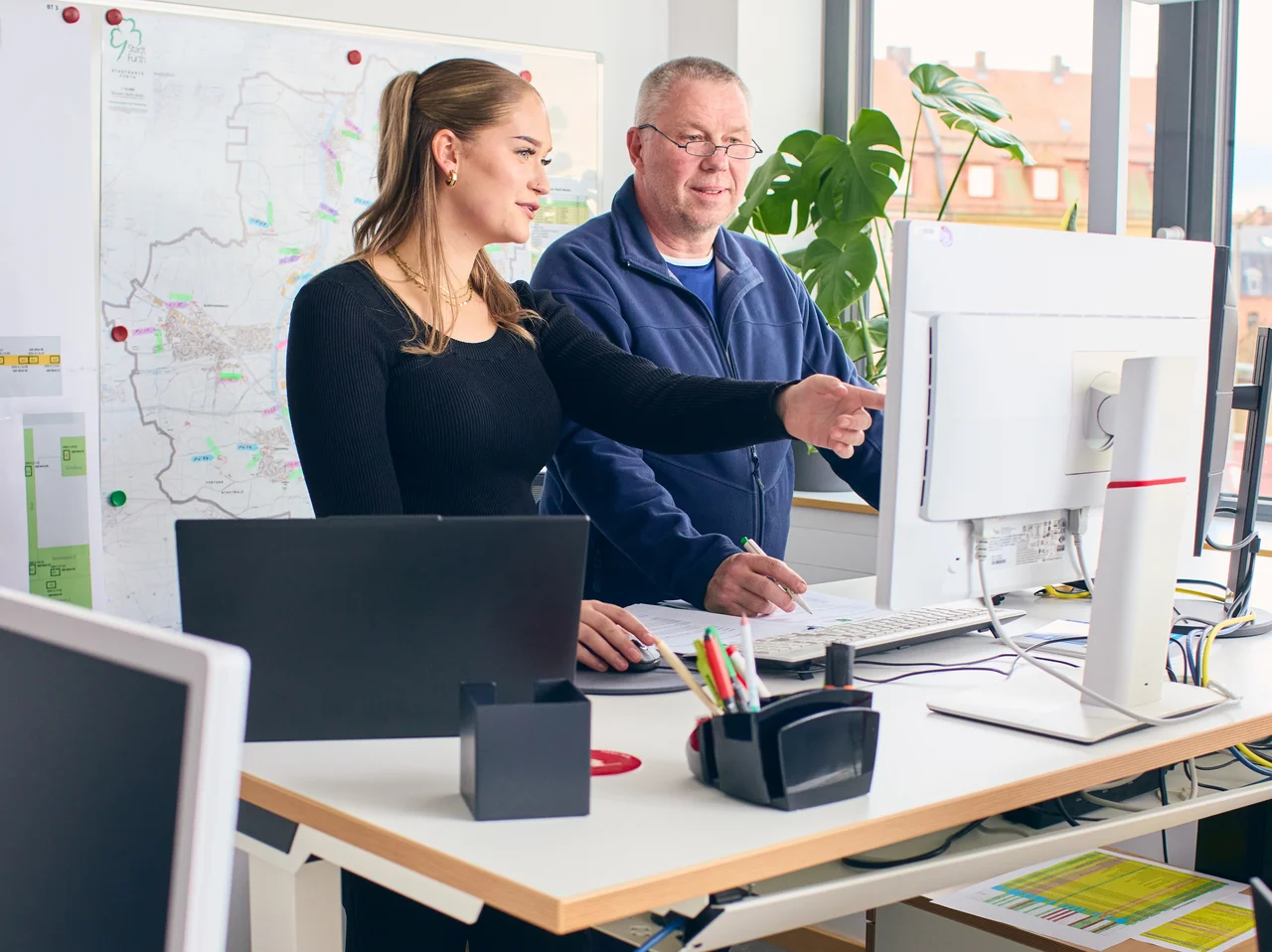 Zwei Personen stehen an einem Schreibtisch und schauen auf einen Computerbildschirm in einem Büro mit mehreren Monitoren.