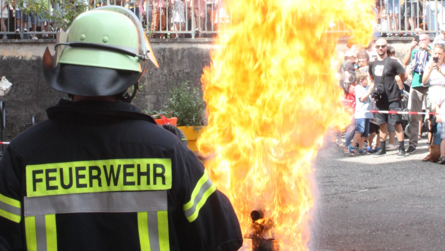 Feuerwehrmann mit Helm und Schutzkleidung steht vor einer großen Flammenwand, im Hintergrund Zuschauer hinter Absperrung