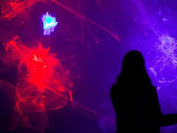 Ausstellungsansicht. Wabernde Lichtwellen in Rot, Blau und Lila. Davor die schwarze Silhouette einer Frau.