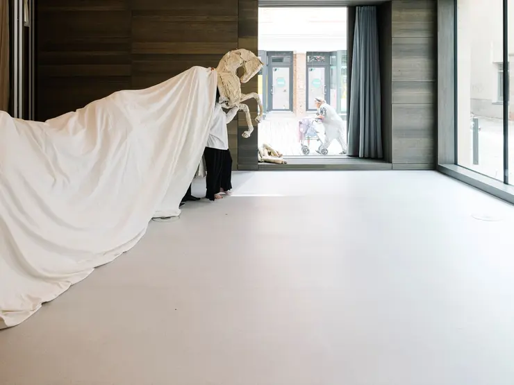 Eine Person in einem weißen Outfit zieht eine große, weiße Stoffhülle, die eine Skulptur oder Figur verdeckt, in einem modernen Raum mit großen Fenstern.