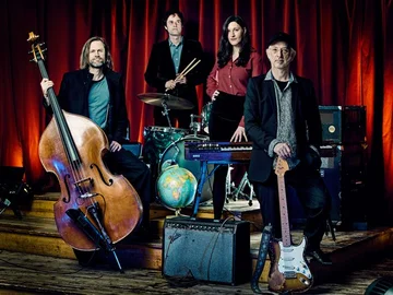 Die band mit Musikinstrumenten vor rotem Vorhang, darunter ein Kontrabass, Schlagzeug, E-Gitarre und ein Verstärker