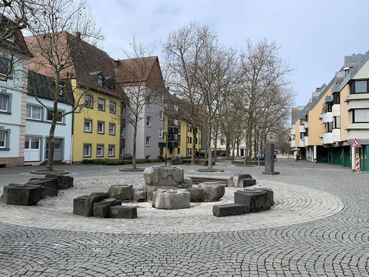 Löwenplatz mit Brunnen im Vordergrund sowie Bäumen und Häusern im Hintergrund. Klick öffnet die Meldung.