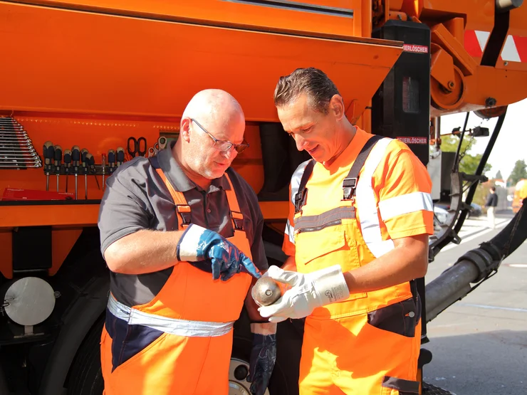 Zwei Mitarbeiter, die an einem Hochdruckspülfahrzeug stehen, ein Objekt halten und gemeinsam betrachten. 