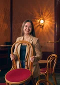Frau in beiger Jacke hält einen Stuhl mit rotem Sitzpolster in einem klassisch eingerichteten Raum mit Holzstühlen und Wandlampen.