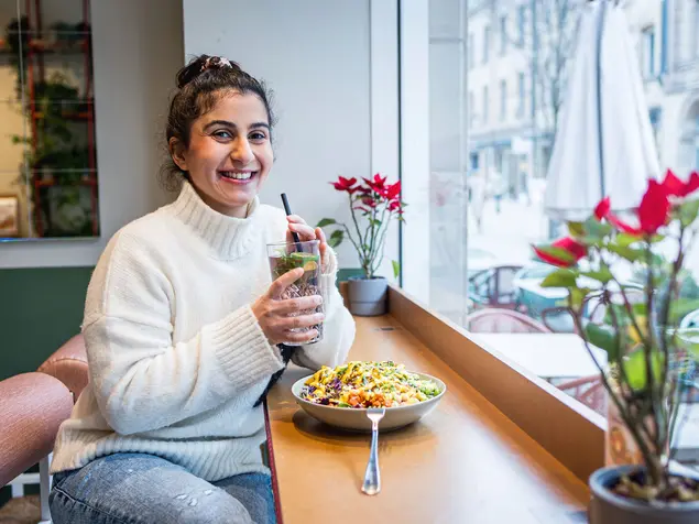 Im Schaufenster eines Cafe mit Blick auf die Straße genießt eine junge Frau einen bunten Salat und ein frisches Getränk.