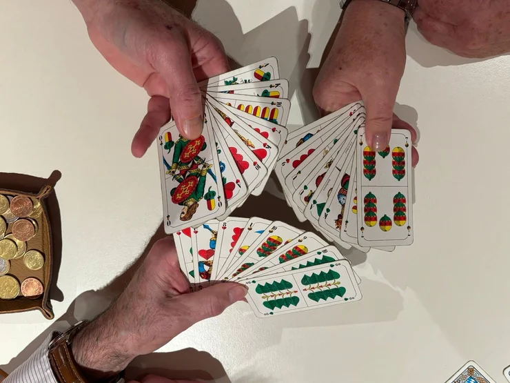 Zwei Hände halten und fächern Spielkarten mit traditionellen deutschen Symbolen über einem weißen Tisch auf, daneben eine Schale mit Münzen