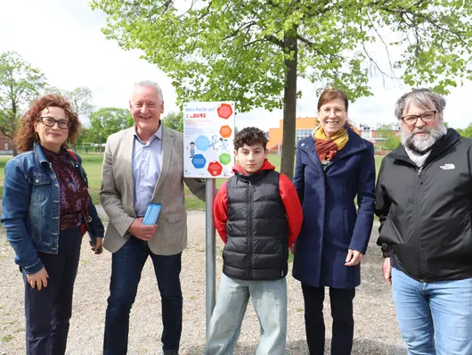 Die Teilnehmenden der Pfaderöffnung präsentieren das Schild zum Recht auf Bildung, im Hintergrund ist die grüne Halle zu sehen. Klick öffnet Bild in größerer Ansicht.