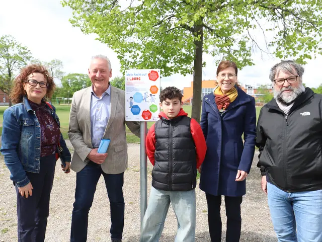 Die Teilnehmenden der Pfaderöffnung präsentieren das Schild zum Recht auf Bildung, im Hintergrund ist die grüne Halle zu sehen. Klick öffnet Bild in größerer Ansicht.