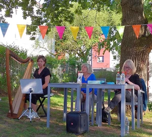 Drei Frauen sitzen an einem Tisch im Freien unter einem Baum, eine Frau spielt eine Harfe, bunte Wimpelketten hängen darüber.