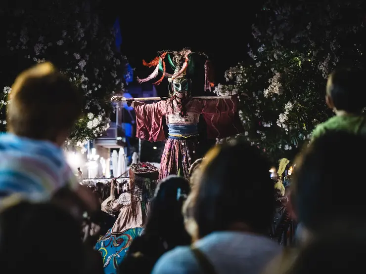 Eine Person in traditioneller Kleidung mit einer auffälligen Maske steht bei Nacht auf einer Bühne, umgeben von Zuschauer*innen, darunter ein Kind auf den Schultern der Eltern.