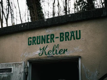 Außenbeschriftung mit grünem Schriftzug 'GRÜNER-BRÄU Keller' über einem offenen Eingang an einer hellen Wand.