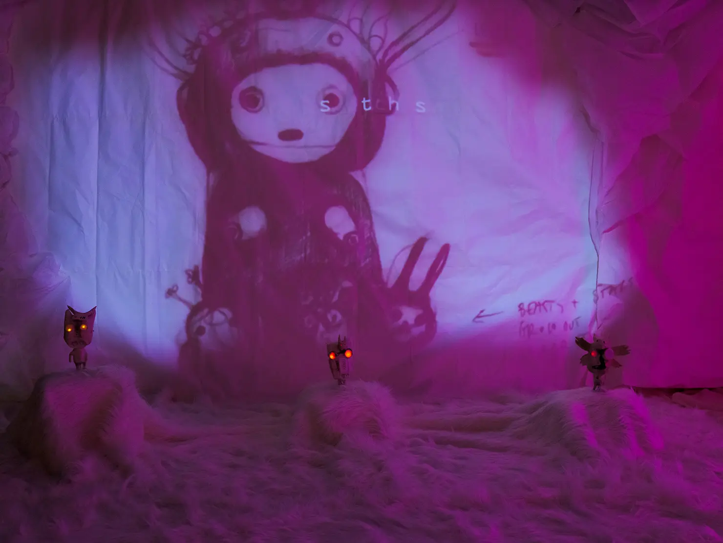 Bühnenbild mit einer Projektion eines fantasievollen, grafisch gestalteten Charakters auf einer weißen Leinwand, umgeben von kreativ gestalteten Objekten im Vordergrund, beleuchtet von rosa und lila Licht.