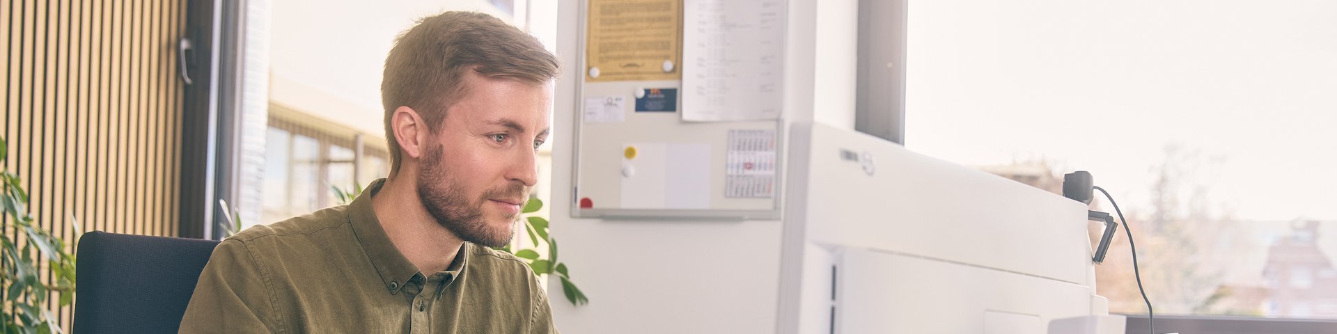 Junger Mann sitzt am Schreibtisch vor einem Computer und hält einen grünen Ordner in der Hand.