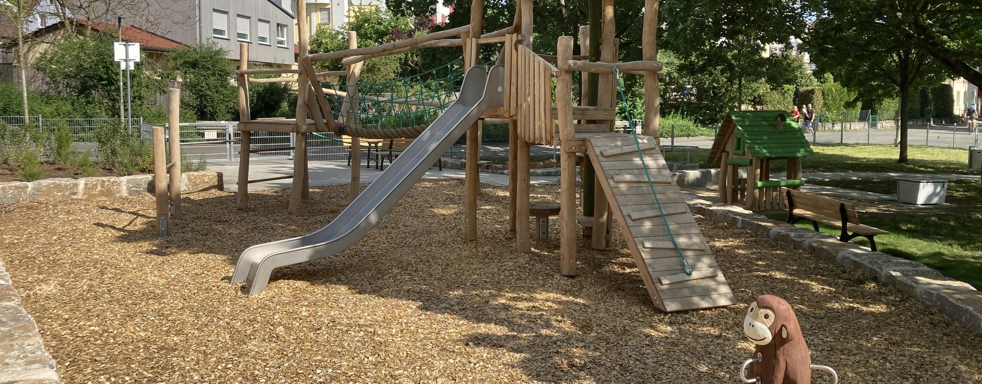 Unter altem Baumbestand steht ein großes Kombinationsspielgerät mit Rutsche und Klettermöglichkeiten. Im Hintergrund Bänke und Tische.