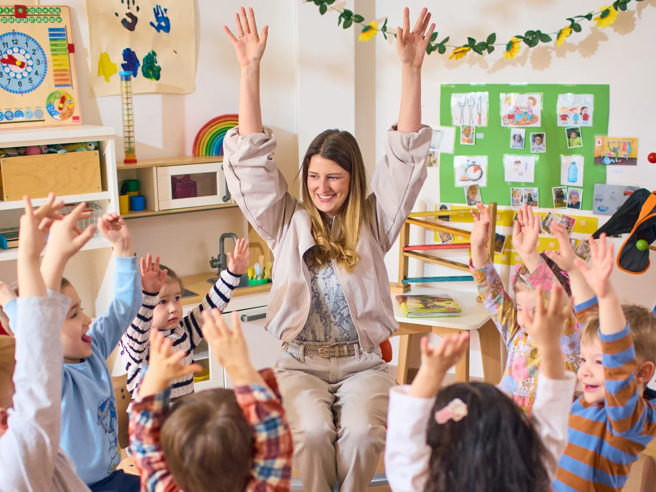 Frau sitzt im Stuhlkreis mit mehreren Kindern im Kindergarten, alle heben die Arme nach oben.