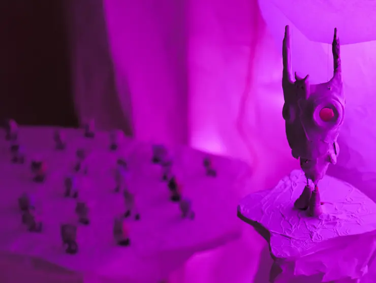 Skulptur mit spitzen Ohren auf einem Podest, umgeben von weiteren kleinen Figuren, beleuchtet in pinkem Licht.