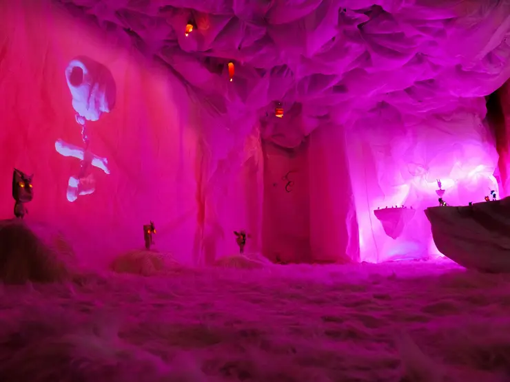 Futuristische Innenansicht eines Raumes mit rosa beleuchteten Wänden und einer Nebelartigen Bodenfläche, dekoriert mit schwebenden Lampen und Projektionen.