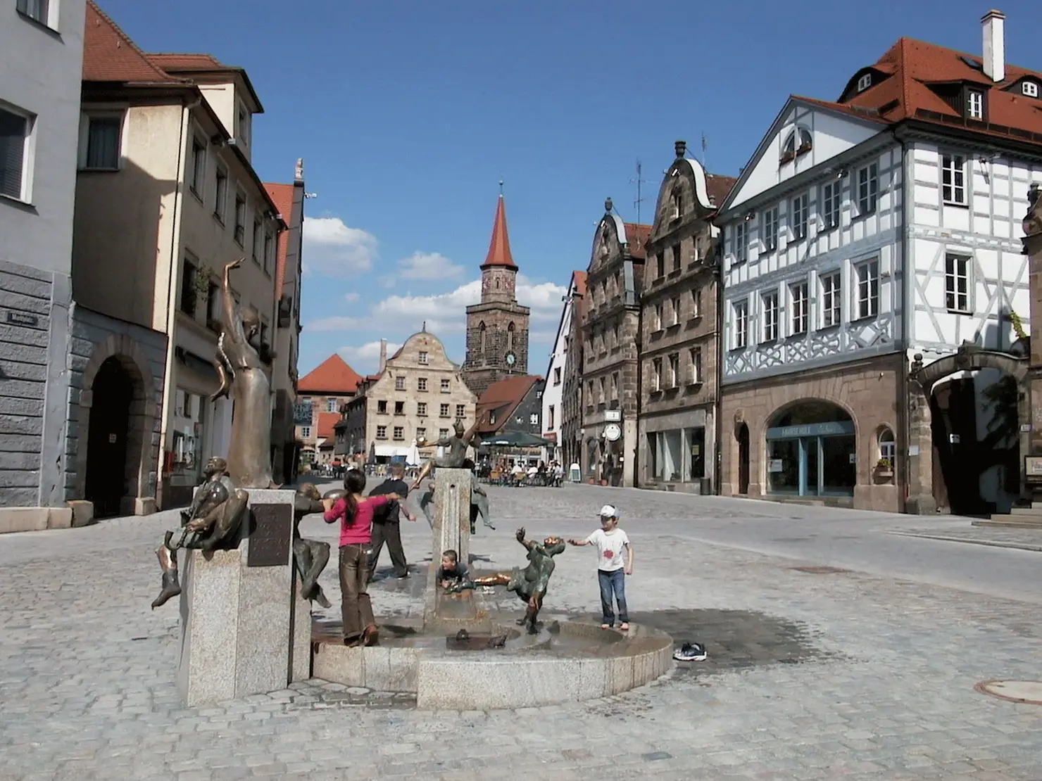 Blick auf den grünen Markt mit Gauklerbrunnen und spielenden Kindern. Im Hintergrund der Kirchturm von St. Michael.