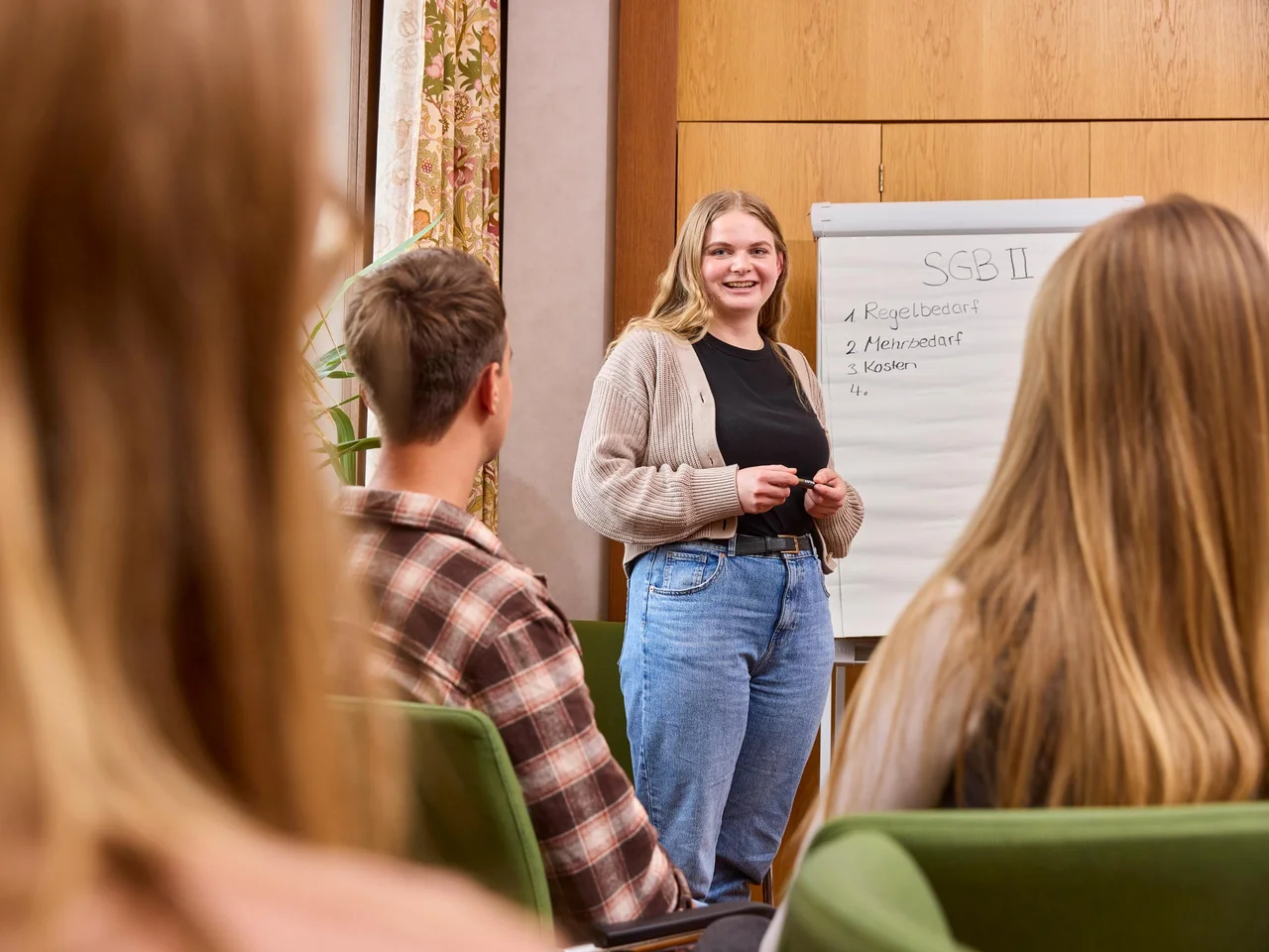 Eine junge Frau steht vor einem Flipchart spricht zu einer Gruppe.