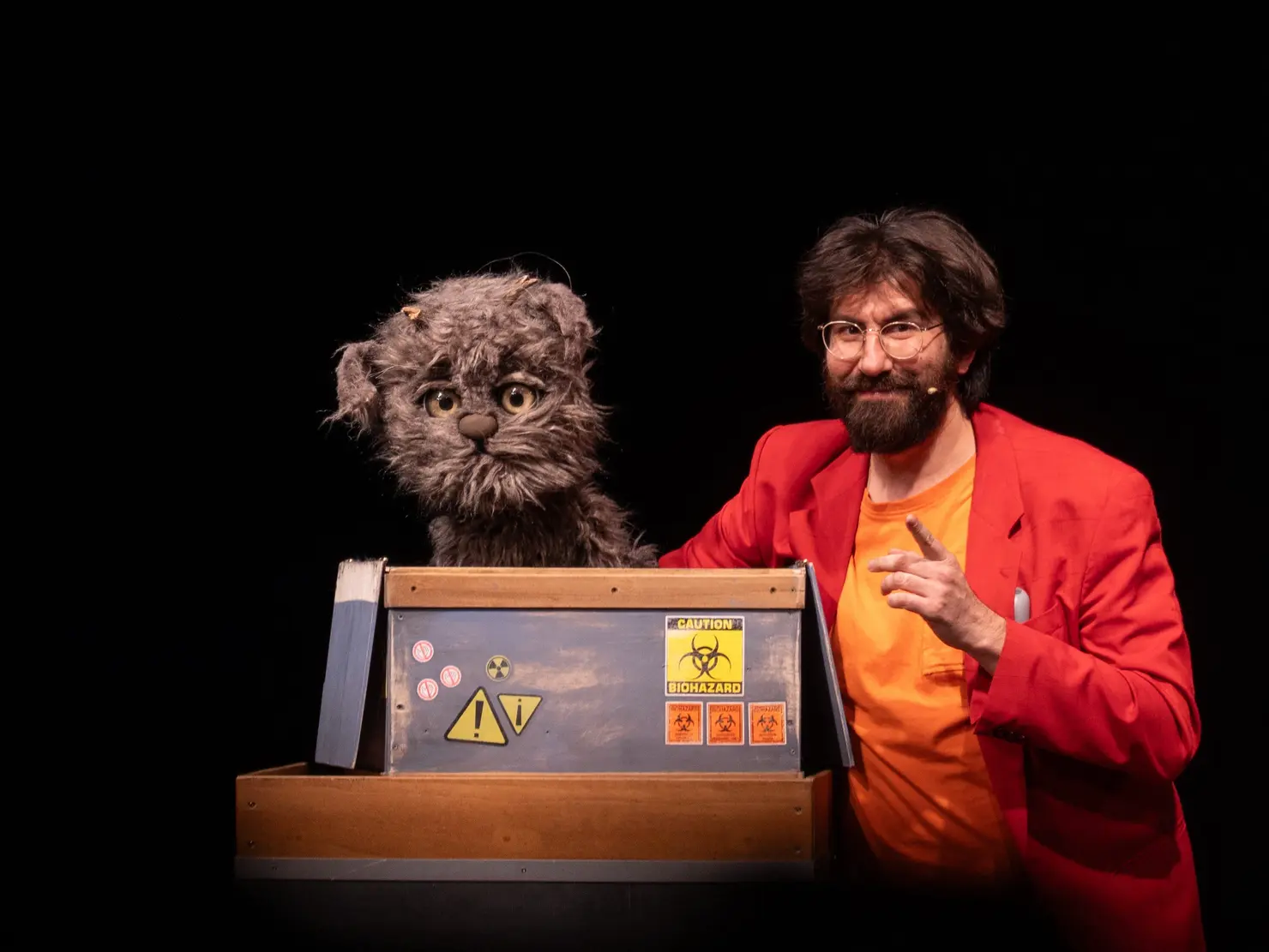 Mann mit Brille und Bart in rotem Sakko steht neben einer katzenähnlichen Stoffpuppe mit grauem Fell und großen Augen, die aus einer Kiste mit Warnsymbolen, die auf eine Giftgefahr hinweisen, herausschaut.