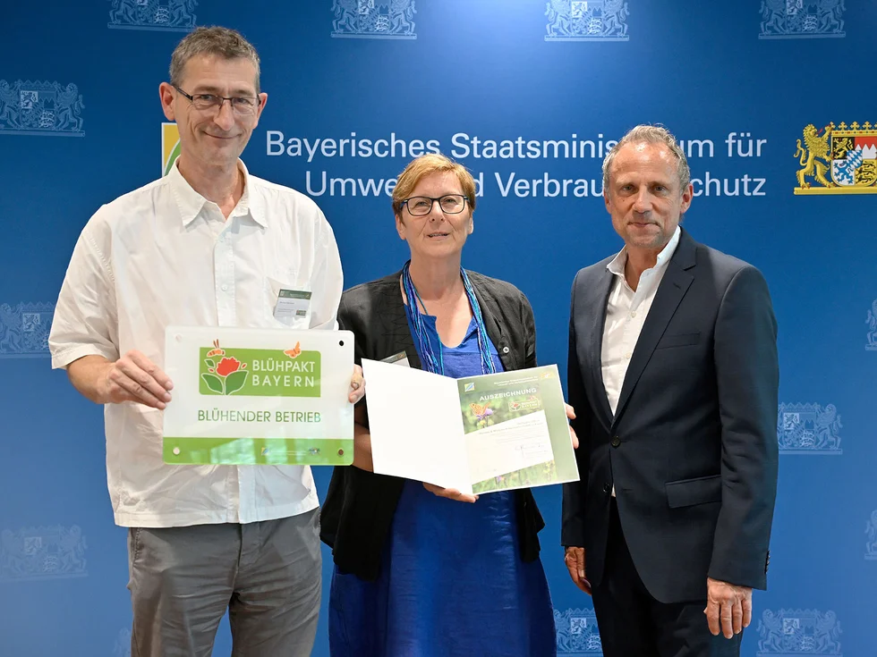 Michael und Renate Killermann stehen mit Umweltminister Thorsten Glauber vor einer blauen Wand mit dem Schriftzug 'Bayerisches Staatsministerium für Umwelt und Verbraucherschutz', sie halten ein Schild mit dem Text 'Blühpakt Bayern Blühender Betrieb' und eine Mappe mit Dokumenten