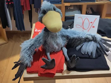Handpuppe in Form eines Vogels sitzt auf gefalteter Kleidung auf einem Holztisch in einem Geschäft, daneben ein Schild mit roter Aufschrift '20%'