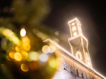 Das festlich beleuchtete Rathaus mit hervorgehobenem Turm, unscharf im Vordergrund Weihnachtsbaumkugeln mit Lichtern.