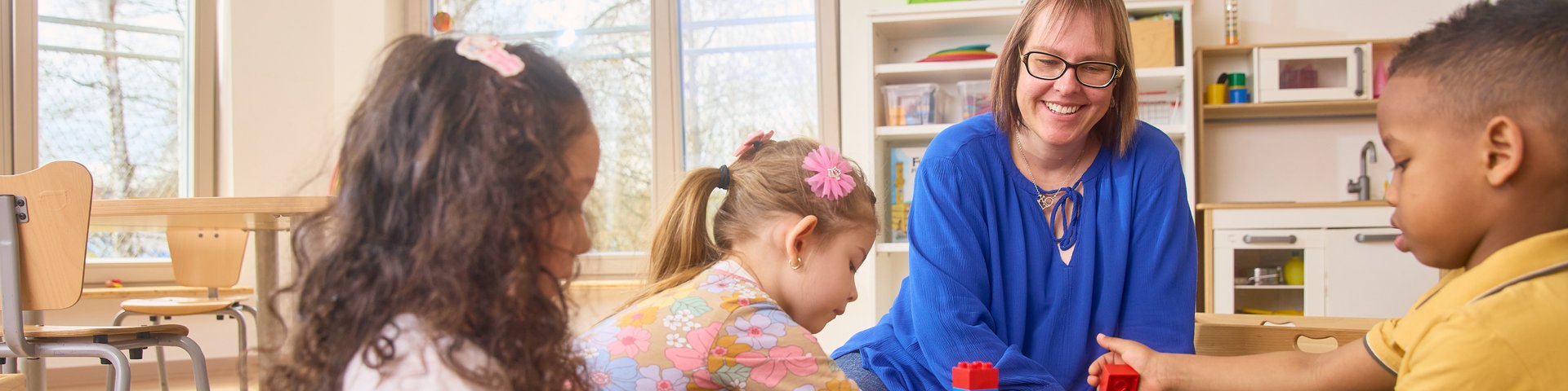Drei Kinder und eine Frau sitzen auf dem Boden und bauen mit bunten Bausteinen in einem Raum mit Kinderzeichnungen an der Wand.