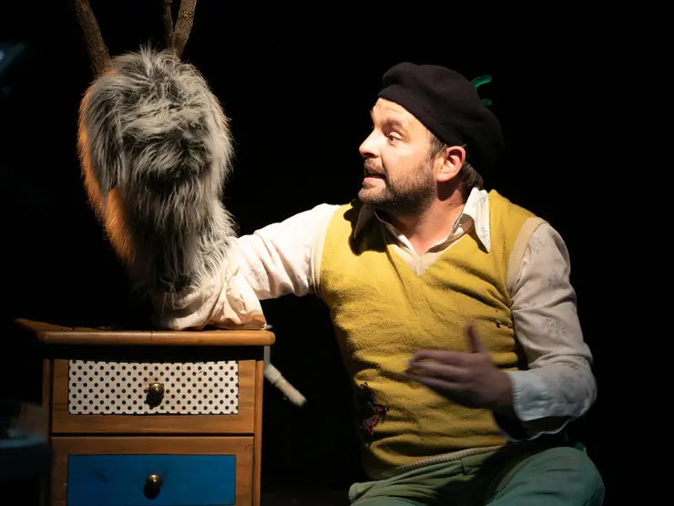 Schauspieler in gelbem Pullover und schwarzem Hut, sitzt neben einem Holzschrank mit einer Handpuppe und gestikuliert während einer Aufführung.