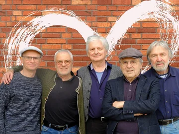 Die fünf Musiker von Young at heart stehen vor einer roten Backsteinwand, alle lächeln und haben die Arme umeinander gelegt.